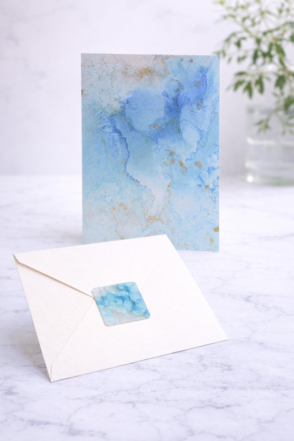 Greeting card with blue watercolor design, matching sticker, and envelope on a marble surface.